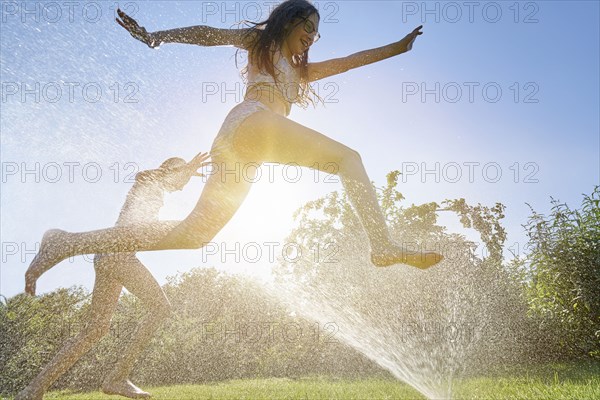 Caucasian girls running and jumping through backyard sprinkler