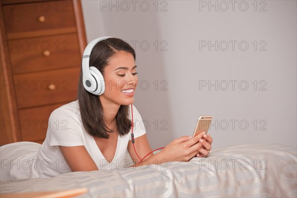 Hispanic woman laying on bed listening to cell phone with headphones
