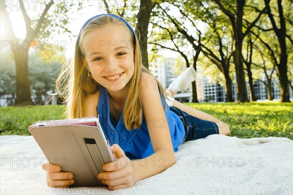 Smiling Caucasian girl laying on blanket in park reading digital tablet