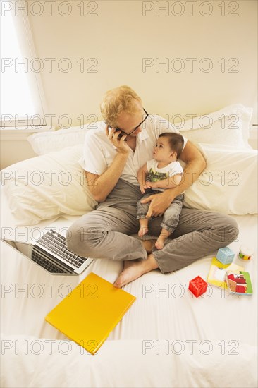 Father holding son and talking on cell phone on bed