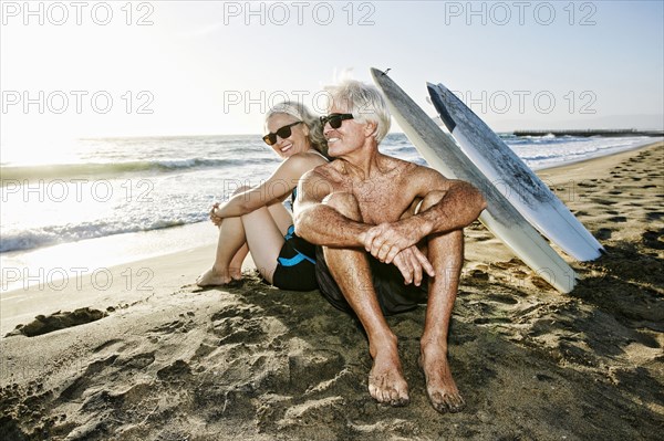 Older Caucasian couple sitting on beach with surfboards