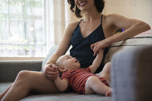 Caucasian mother sitting on sofa holding hands of baby son