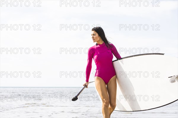 Woman in pink swimsuit carrying paddleboard by lake