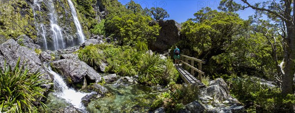 Rear view of hiker crossing footbridge near waterfall in Fiordland National Park, Te Anau, Fiordland, New Zealand