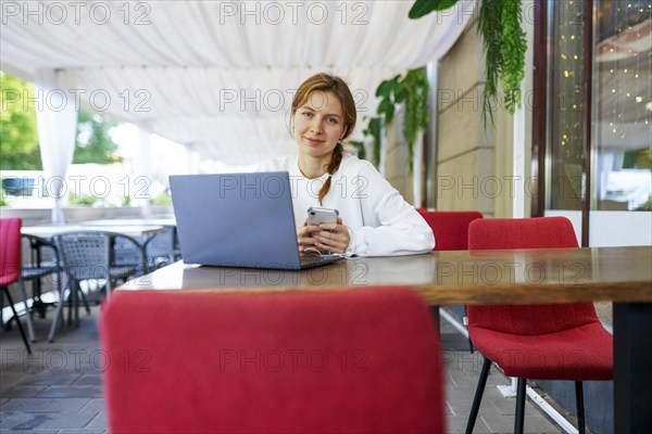 Portrait of smiling woman with smart phone and laptop at cafe table, Omsk, , Russia