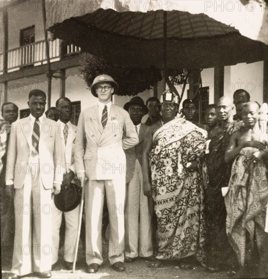 Empire Day in Aburi, 1950. A European colonial officer stands beneath a shady parasol with an Agona chief, posing for a portrait during celebrations for Empire Day. A group of traditionally dressed Agona men and women, and several African men dressed in Western-style clothing surround the two men. Aburi, Gold Coast (Ghana), 24 May 1950. Aburi, East (Ghana), Ghana, Western Africa, Africa.