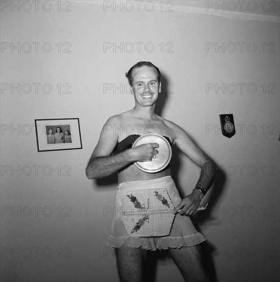 Fan handler. Ron Richardson performing a fan dance at the Archer's New Year party wearing only a maid's apron and bra. He holds a pan lid up in one hand and a wicker fan in the other. Kenya, 31 December 1954. Kenya, Eastern Africa, Africa.