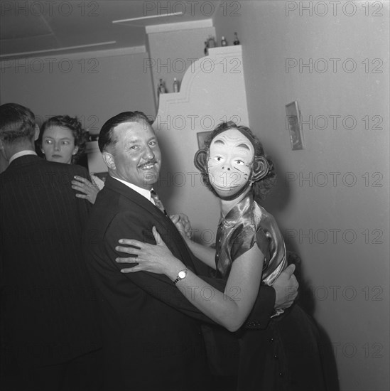 Monkeying around. Couples dancing at the Archer's New Year party. The woman in the foreground wears a child's animal mask as she dances with her partner. Kenya, 31 December 1954. The man pictured is wearing a fancy dress baby costume and holds a bottle of Crawfords old scotch whiskey in one hand. The small crowd of onlookers in the background are clearly entertained. Kenya. 31 December 1954, Kenya, Eastern Africa, Africa.