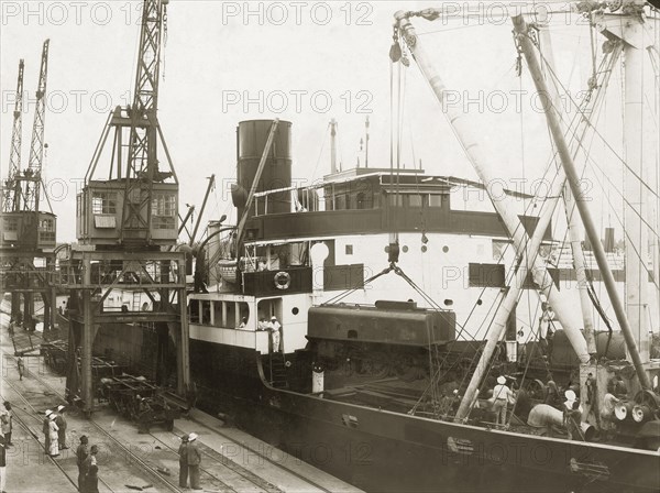 Unloading a locomotive tender from SS Harmonides. Cranes onboard the SS Harmonides hoist a locomotive tender (water tank railcar or wagon) above the ship's decks at Kilindini harbour. Mombasa, Kenya, circa 1926. Mombasa, Coast, Kenya, Eastern Africa, Africa.