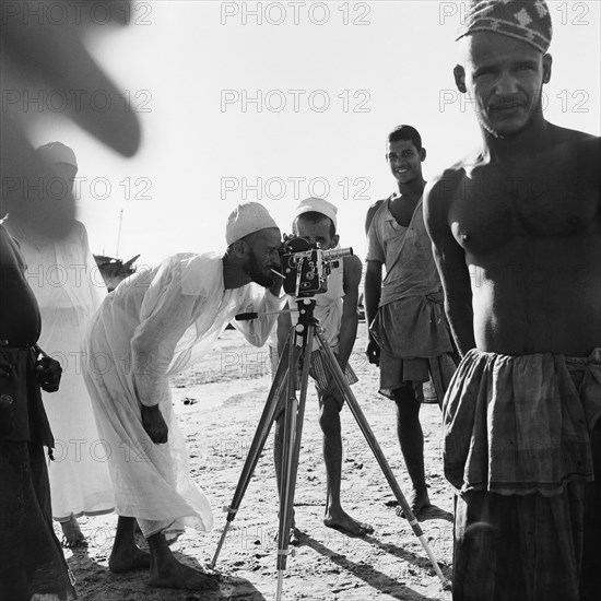 Amateur photographer. A man smoking a cigarette bends over to peer into the viewfinder of a Paillard-Bolex movie camera mounted on a tripod. A group of casually dressed, barefooted men surround him, two of them looking directly into Trotter's lens. Zanzibar (Tanzania), 3-12 March 1953., Zanzibar Central/South, Tanzania, Eastern Africa, Africa.
