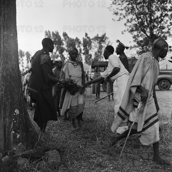 Cleansing ceremony. Two men brush bundles of branches against a woman wearing jewellery as she passes between them at a cleansing ceremony. Another man holding a baton shouts at a line of people queuing for the same treatment. Kenya, 20-28 March 1953. Kenya, Eastern Africa, Africa.