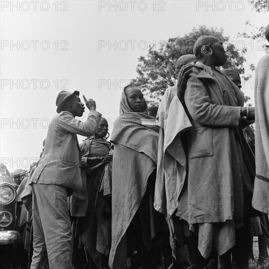 Queuing at a cleansing ceremony. A man with a raised finger speaks over the top of a crowd of women queuing for a cleansing ceremony. Kenya, 20-28 March 1953. Kenya, Eastern Africa, Africa.