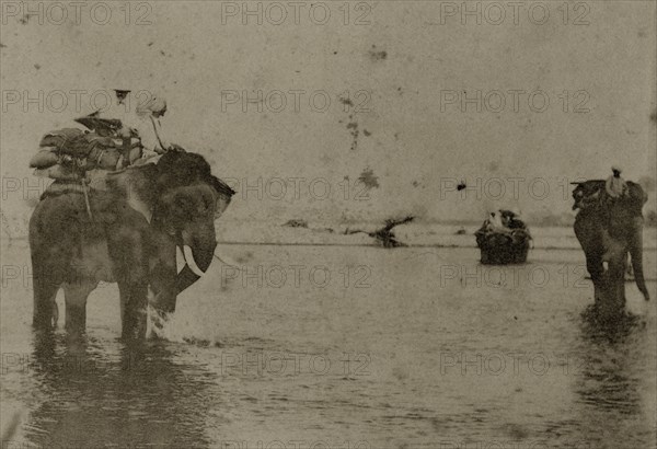 Hunting elephants wet their feet. Two hunting elephants, laden with blankets, cool their feet in a shallow river, their mahouts (elephant handlers) sitting patiently on their shoulders. North East India, circa 1890. India, Southern Asia, Asia.