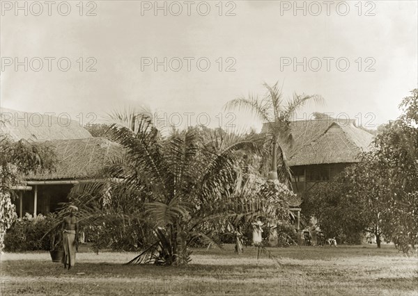 In the garden of a colonial bungalow. A barechested servant carries a bucket across the lawn of a colonial bungalow: its British inhabitants relax in deckchairs to the right. Burma (Myanmar), circa 1946. Burma (Myanmar), South East Asia, Asia.