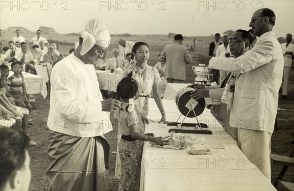 Raffle for six bottles of whisky. The President of the Rangoon Golf Club spins a drum full of raffle tickets in front of an audience, ready to be drawn by two young Burmese girls. Rangoon (Yangon), Burma (Myanmar), circa 1952. Onlookers wait in anticipation for the results of the raffle at the Burma open Championship. Rangoon, Burma, 1952. Yangon, Yangon, Burma (Myanmar), South East Asia, Asia.