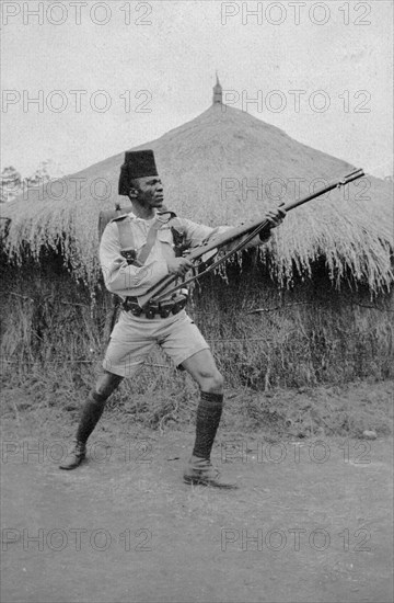 Askari from Second King's African Rifles. An African askari (soldier) poses with his gun in front of a grass-roofed dwelling. A caption identifies him as a Guard Sergeant Major of the Second Battalion of the King's African Rifles. British East Africa (Kenya), circa 1916. Kenya, Eastern Africa, Africa.