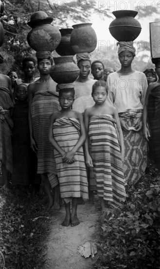 Carrying pots. Several Nigerian women and a young girl wear wraparound blankets and balance large clay pots on their heads in the midst of a crowd that has gathered for the photograph. Ifaki-Ekikti, Nigeria, circa 1935. Ifaki, Ekiti, Nigeria, Western Africa, Africa.