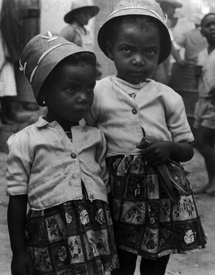 Grenadian schoolgirls. Two small Grenadian schoolgirls stand close together dressed in matching bonnets, buttoned shirts and patterned skirts. The taller of the two holds a bunch of leaves in her hand. Grenada, West Indies, 1965. Grenada, Caribbean, North America .