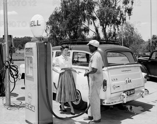 Daphne Dale fills up with Shell. Promotional shot of Daphne Dale, posing for the camera beside a car at a roadside petrol station. An African workman faces her, holding the hose of a Shell petrol pump. Kenya, 11 November 1955. Kenya, Eastern Africa, Africa.