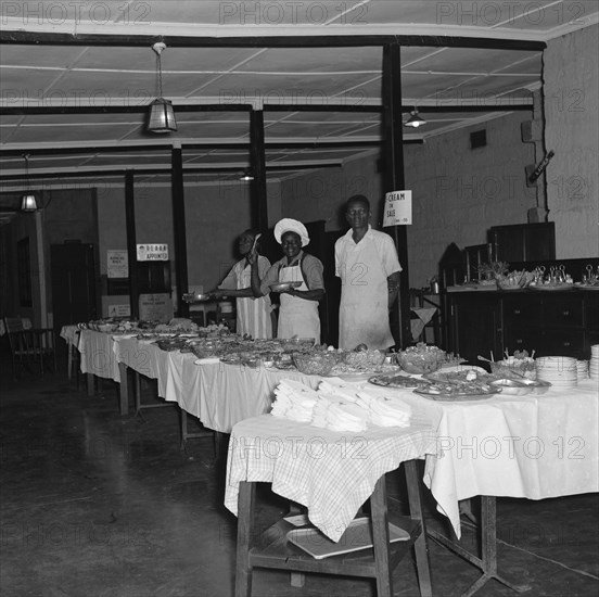 Buffet for the SJAK dance. Three African men wait to serve behind a long line of buffet tables covered with food at a dance held for the Sports Journalists Association of Kenya (SJAK). Kenya, 19 August 1955. Kenya, Eastern Africa, Africa.