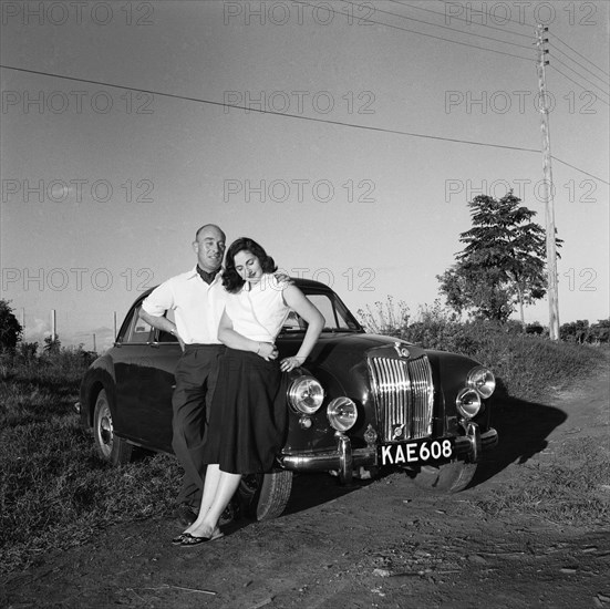 Sue and John Paton. Sue and John Paton relax, leaning back against their MG car in the evening sunlight. She wears a wrapover top and embroidered Chinese-style slippers: he wears a cravat. Kenya, 24 October 1955. Kenya, Eastern Africa, Africa.