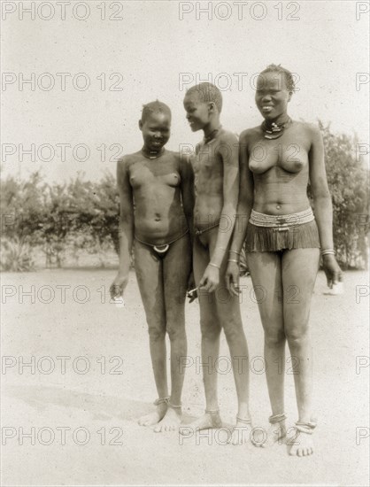 Sudanese youths. Three Sudanese youths stand closely together, naked apart from the minimal jewellery they are wearing. The young woman on the right displays a pattern of scars on her midriff. Southern Sudan, circa 1928. Sudan, Eastern Africa, Africa.