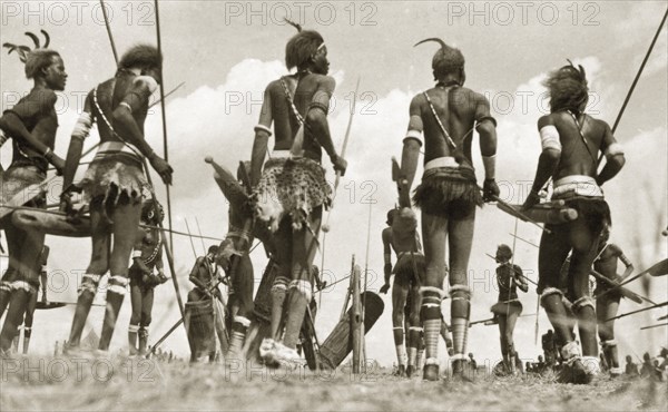 Sudanese warrior costume. A group of Sudanese men gather in a circle holding spears and drums. They wear traditional warrior costume comprising headdresses, body jewellery and animal skins. Sudan, circa 1930. Sudan, Eastern Africa, Africa.