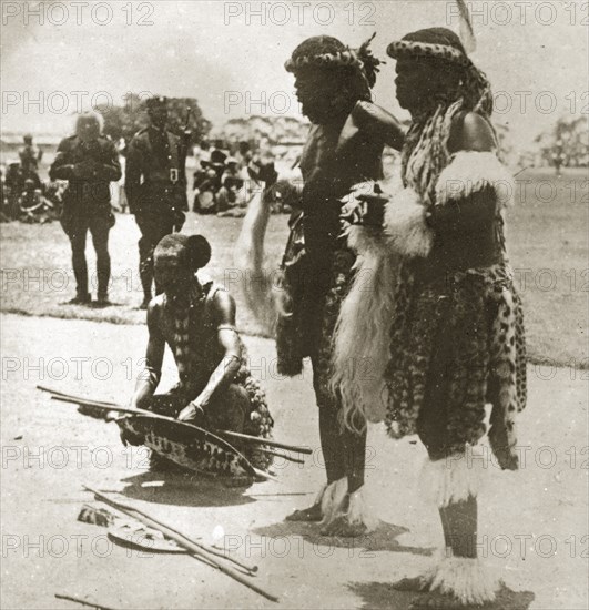 Three Zulu chiefs. Three Zulu chiefs dressed in ceremonial costume made from cheetah fur, gather before an African audience with shields and sticks. Durban, Natal (KwaZulu Natal), South Africa, 1-5 January 1924. Durban, KwaZulu Natal, South Africa, Southern Africa, Africa.