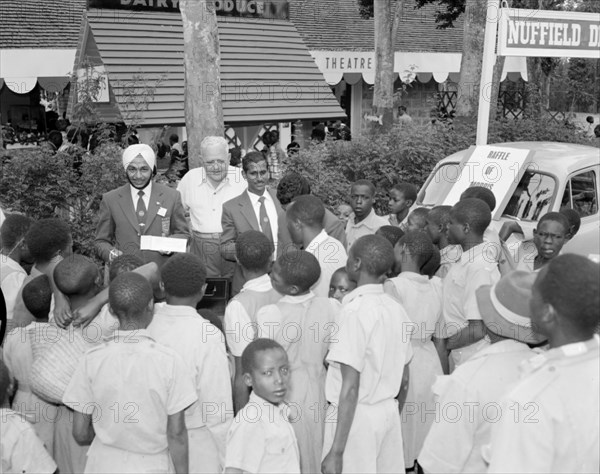 The Olympic Association meets schooldchildren. Two Asian men representing the Olympic Association talk to a group of African schoolchildren who crowd around them at the Royal Show. Kenya, 17-20 October 1956. Kenya, Eastern Africa, Africa.