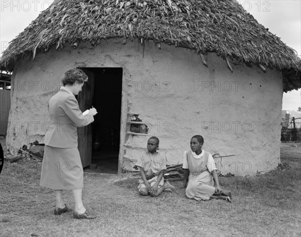 A book artist sketches children. Promotional shot for the East African Literature Bureau. A European artist sketches two African children sitting beside a hut with a thatched roof. Kenya, 25 September 1956. Kenya, Eastern Africa, Africa.