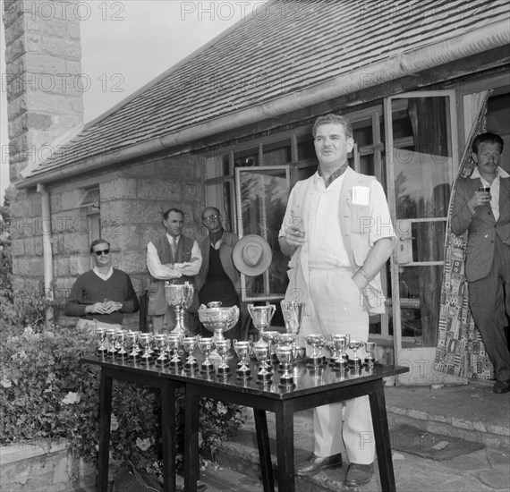 Prize-giving speech. Eric Mathers gives a speech, cigarette in hand, at the prize-giving ceremony of the Brackenhurst Hill Climb. A table of trophies is laid out before him. Limuru, Kenya, 30 September 1956. Limuru, Central (Kenya), Kenya, Eastern Africa, Africa.