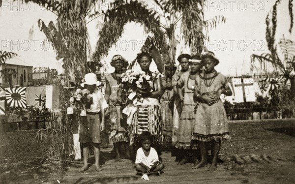 Fijian women. A group of Fijian women and children wearing traditional dress gather beneath an arch of trees. The woman and boy in the centre hold bunches of flowers, others wear flowered headbands. Suva, Fiji, 21-27 May 1924. Suva, Viti Levu, Fiji, Pacific Ocean, Oceania.