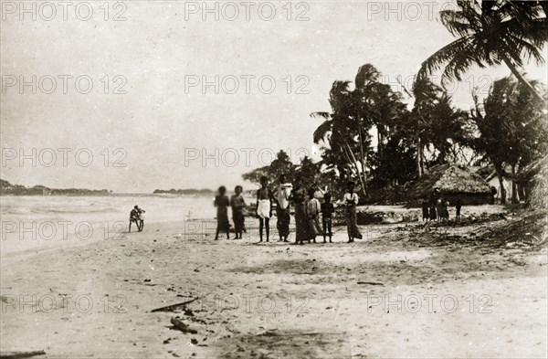 Ceylonian seashore. A group of Ceylonian people stand facing the camera on a beach. Thatched village huts and palm trees are visible in the background. Trincomoli, Ceylon (Sri Lanka), 27-31 January 1924. Trincomali, East (Sri Lanka), Sri Lanka, Southern Asia, Asia.