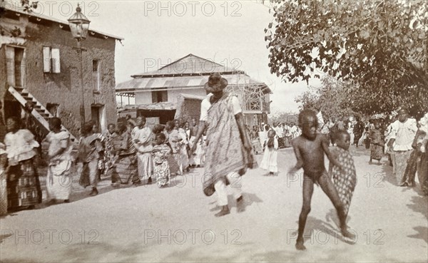 Traditional street dance. A man and children dance between long lines of women who perform a traditional dance along the sides of a street. Possibly Ghana, Africa, circa 1920. Africa.