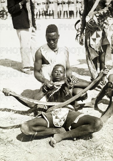 Snake charming, Tanganyika. Snake charming for an audience. The principal perfomer holds onto the head and tail of a snake wrapped tightly around his waist whilst another man wraps a smaller snake around his neck. Tanganyika Territory (Tanzania), circa 1960. Tanzania, Central Africa, Asia.