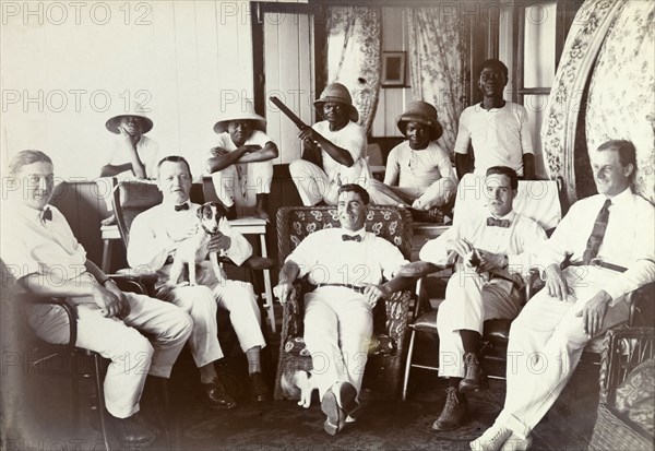 Miller Brothers agency staff relaxing. Informal portrait of the British managers of Miller Brothers agency and their African house servants relaxing at the company bungalow. Young servants are perched on tables behind their European employers. They are wearing their employers' solatopis or pith helmets, and one of them brandishes a gun. Accra, Gold Coast (Ghana), circa 1918. Accra, East (Ghana), Ghana, Western Africa, Africa.