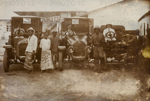 Men and cars, Western Africa. Three European drivers sit behind the wheels of their cars, surrounded by a several African men. Western Africa, circa 1920, Western Africa, Africa.