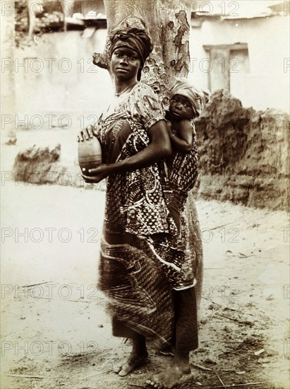 Mother and baby. Outdoors portrait of a young mother with her baby bound to her back. She wears one or more wrap cloths over the top of a tailored blouse with short puffed sleeves. Both she and the baby wear head cloths. Western Africa, circa 1920., Western Africa, Africa.