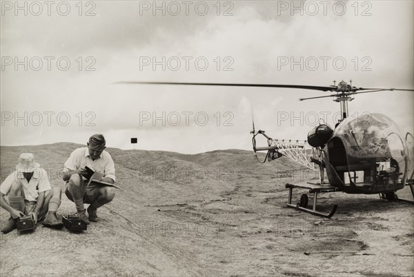 Surveying with a Bell 47G helicopter. Two men crouch down beside a Bell 47G helicopter as they survey the surrounding land from a hilltop using scientific equipment. Probably Southern Rhodesia (Zimbabwe), circa 1955. Zimbabwe, Southern Africa, Africa.