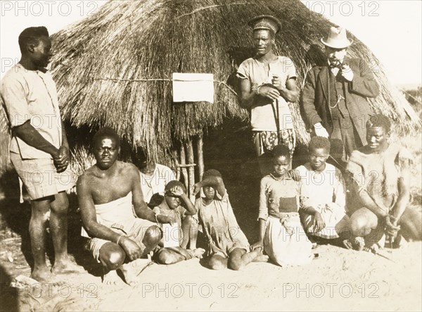 Portrait of workers and children at Balloch Farm. Portrait of a group of male African farm workers, assembled outdoors beside a round, thatched hut at Balloch Farm. They pose with four children and a European man, probably an overseer, who wears a suit and smokes a pipe. Near Gwelo, Southern Rhodesia (near Gweru, Zimbabwe), circa 1930. Gweru, Midlands, Zimbabwe, Southern Africa, Africa.