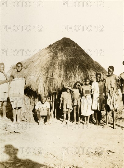 African workers and children at Balloch Farm. Portrait of a group of male African farm workers and their children, lined up in height order outside a round, thatched hut at Balloch Farm. Near Gwelo, Southern Rhodesia (near Gweru, Zimbabwe), circa 1930. Gweru, Midlands, Zimbabwe, Southern Africa, Africa.