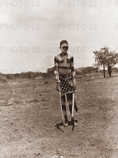 An African dancer at Balloch Farm. Outdoors portrait of an African dancer at Balloch Farm. He holds a long stick in each hand and stands naked from the waist up, his body and face daubed with pigment. Near Gwelo, Southern Rhodesia (Gweru, Zimbabwe), circa 1930. Gweru, Midlands, Zimbabwe, Southern Africa, Africa.