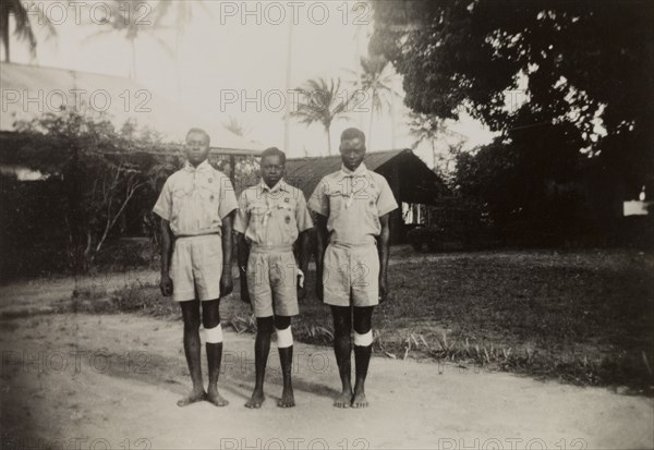 Three Kenyan scouts. Three Kenyan scouts pose for the camera outdoors at an inspection of scouts and guides by Sir Charles Mortimer, the Commissioner for Lands. Each boy wears scout uniform and what appears to be a bandage around his left knee. Coast Province, Kenya, circa 1950., Coast, Kenya, Eastern Africa, Africa.