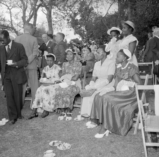 Guests at the Royal Garden Party. A group of African women dressed in Western clothing sit together at the Royal Garden Party. In the background, European and African guests socialise, drinking cups of tea on the lawn. Kenya, 20 October 1956. Kenya, Eastern Africa, Africa.