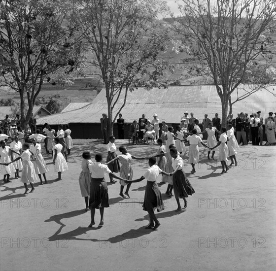 Schoolgirls dance for Princess Margaret. Princess Margaret and her entourage watch a group of African schoolgirls perform a welcoming dance from the shade of trees in the grounds of a school. Machakos, Kenya, 22 October 1956. Machakos, East (Kenya), Kenya, Eastern Africa, Africa.