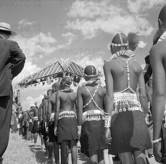 Wakamba costume from behind. Wakamba dancers stand in line with their backs to the camera during Princess Margaret's visit. Their ceremonial dress consists of elaborately beaded necklaces, headbands and belts. Machakos, Kenya, 22 October 1956. Machakos, East (Kenya), Kenya, Eastern Africa, Africa.