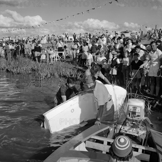 Landing stage at the Aquasports Regatta. Crowds at the Aquasports Regatta watch as three men stand knee-deep in water at the edge of a lake, struggling to control two small motor boats. Kenya, 28 October 1956. Kenya, Eastern Africa, Africa.