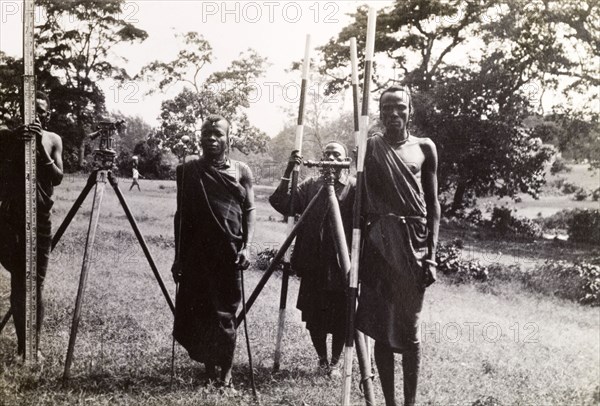 Maasai men help a surveying team. Four Maasai men in traditional dress help a surveying team by holding up distance markers for a hidden photographer. Probably Tanganyika Territory (Tanzania), circa 1930. Tanzania, Eastern Africa, Africa.