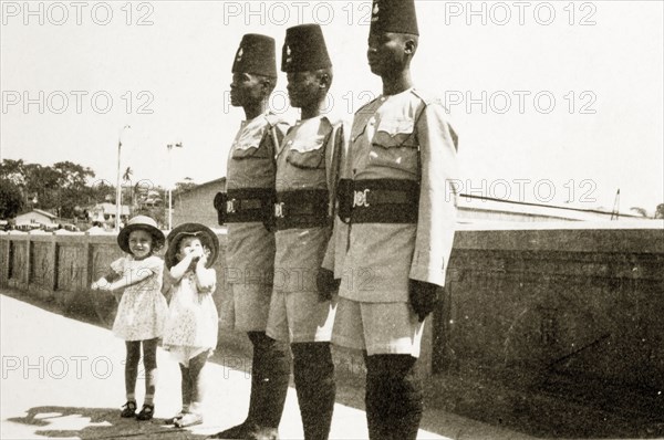 Barbara and Gwynneth with askaris. Two small European girls, Barabara and Gwynneth Wallhouse, pose awkwardly beside three uniformed African askaris (soldier) who stand to attention. Probably Tanganyika Territory (Tanzania), circa 1935. Tanzania, Eastern Africa, Africa.