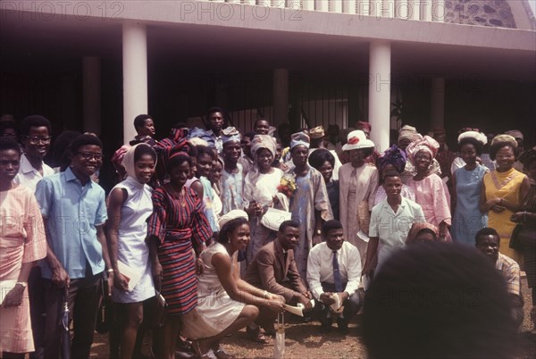 Nigerian wedding. A group of smiling wedding guests, mostly dressed in European-style clothing, squeeze together for a group photograph with the bride and groom outside a university building. Nigeria, circa 1970. Nigeria, Western Africa, Africa.